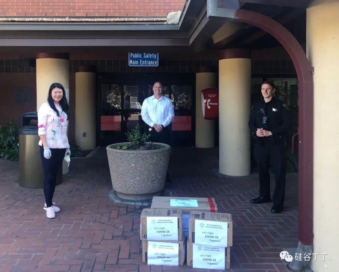 Chamber representatives hand over boxes of masks labeled "Let's Fight COVID-19 Together!" to city staff and a police officer outside a Silicon Valley public safety building, 2020.